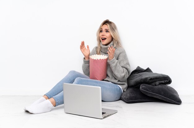 Donna bionda dell'adolescente che mangia popcorn mentre guardando un film sul computer portatile con espressione facciale di sorpresa