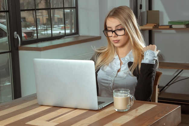 Donna bionda di affari in vetri che lavorano con il computer portatile e beve il caffè