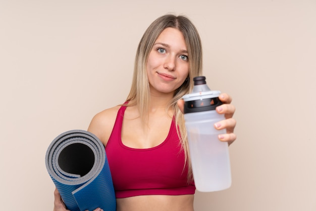 Donna bionda di giovane sport sopra la parete isolata con la bottiglia di acqua di sport e con una stuoia