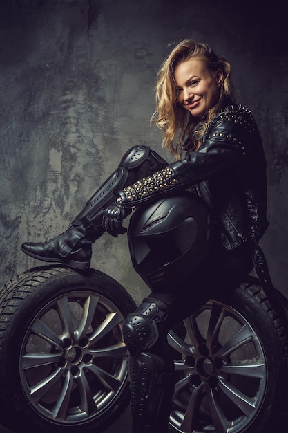 Donna bionda di lusso in uniforme da motociclista in pelle in posa su ruote di auto in studio.