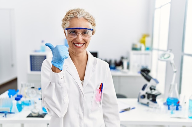 Donna bionda di mezza età che lavora al laboratorio dello scienziato facendo il pollice felice in su gesto con la mano che approva l'espressione guardando la telecamera che mostra successo