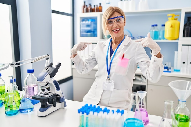 Donna bionda di mezza età che lavora in un laboratorio scientifico con un sorriso sul viso che si indica con le dita orgogliosa e felice