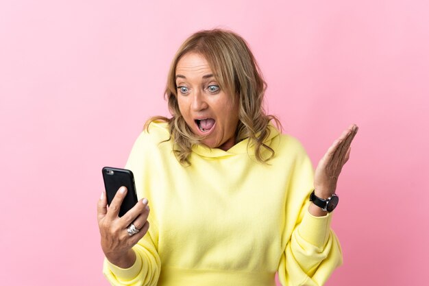 Donna bionda di mezza età sopra il rosa isolato guardando la telecamera mentre si utilizza il cellulare con espressione sorpresa