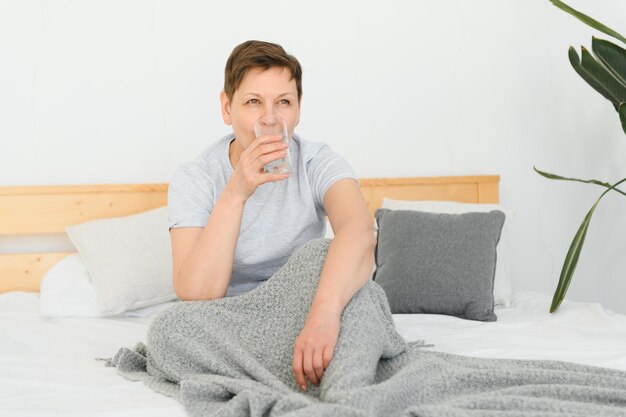 Donna bionda di mezza età sorridente felice bicchiere d'acqua sdraiato sul letto a casa