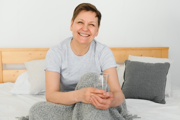 Donna bionda di mezza età sorridente felice bicchiere d'acqua sdraiato sul letto a casa