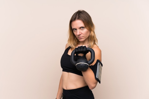 Donna bionda di sport sopra la parete isolata che fa sollevamento pesi con kettlebell e che guarda alla parte anteriore