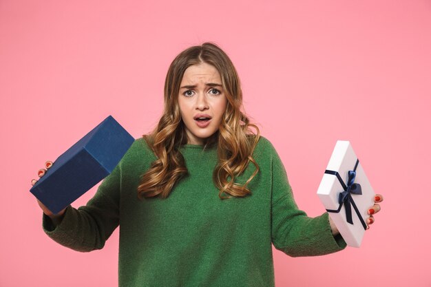 Donna bionda dispiaciuta che indossa un maglione verde che apre una scatola regalo vuota e guarda la parte anteriore sul muro rosa