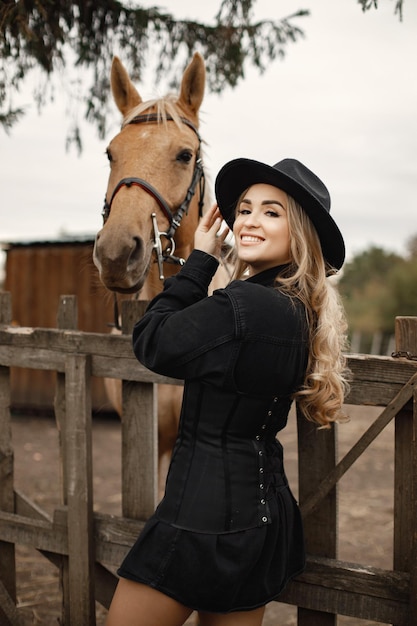 Donna bionda e cavallo marrone in piedi in una fattoria. Donna che indossa abiti neri e cappello. Donna che tocca il cavallo dietro il recinto.