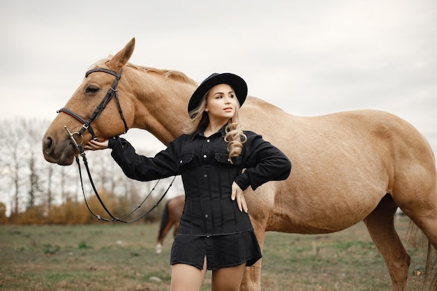 Donna bionda e cavallo marrone in piedi nel campo. Donna che indossa abiti neri e cappello. Donna che tocca il cavallo.