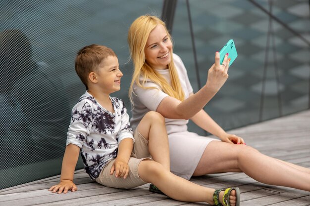 Donna bionda felice e ragazzino seduti sulla terrazza e facendo selfie sullo smartphone Madre e figlio si divertono insieme Giovane mamma positiva che trascorre del tempo con il suo bambino carino Divertirsi Famiglia