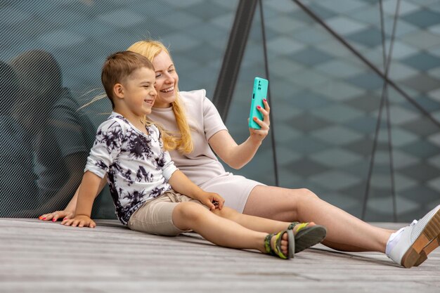 Donna bionda felice e ragazzino seduti sulla terrazza e facendo selfie sullo smartphone Madre e figlio si divertono insieme Giovane mamma positiva che trascorre del tempo con il suo bambino carino Divertirsi Famiglia