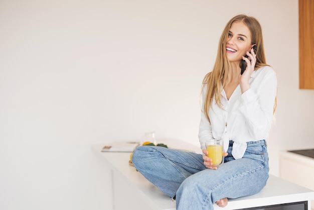 Donna bionda felice e sorridente seduta sopra il tavolo della cucina che beve succo d'arancia e parla al telefono