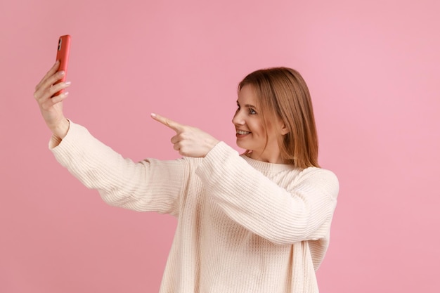 Donna bionda felice sorridente che punta alla fotocamera del cellulare mentre si effettua una videochiamata o un live streaming