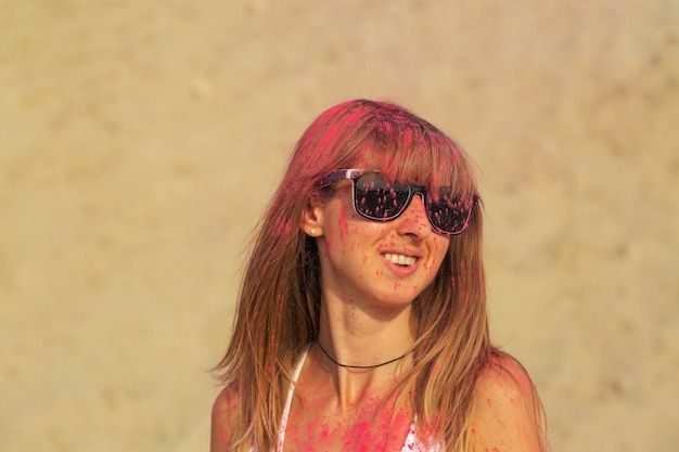 Donna bionda fresca in occhiali da sole ricoperti di vernice secca rosa Holi nel deserto. Spazio vuoto