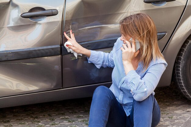 Donna bionda in abbigliamento casual seduta vicino a un'auto ammaccata e toccando graffi e ammaccature sulla portiera dell'automobile, parlando al telefono con l'agente assicurativo o chiedendo aiuto, incidente stradale. Tiro all'aperto.