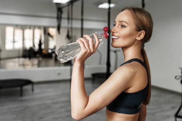 Donna bionda in abbigliamento sportivo che beve acqua dopo l'allenamento in una moderna palestra