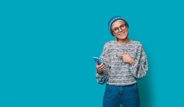 Donna bionda in abiti blu che punta al suo nuovo telefono con gli occhiali da vista su una parete di studio con spazio libero