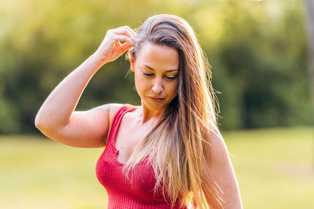 Donna bionda in abiti estivi che si tocca i capelli in un parco con la luce del sole che le illumina i capelli