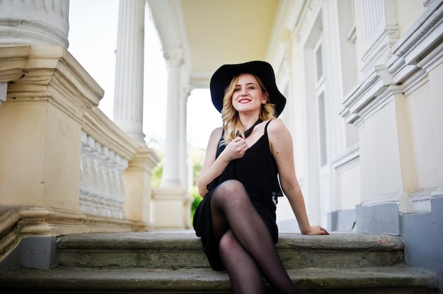 Donna bionda in abito nero, collane e cappello contro la casa d'epoca.