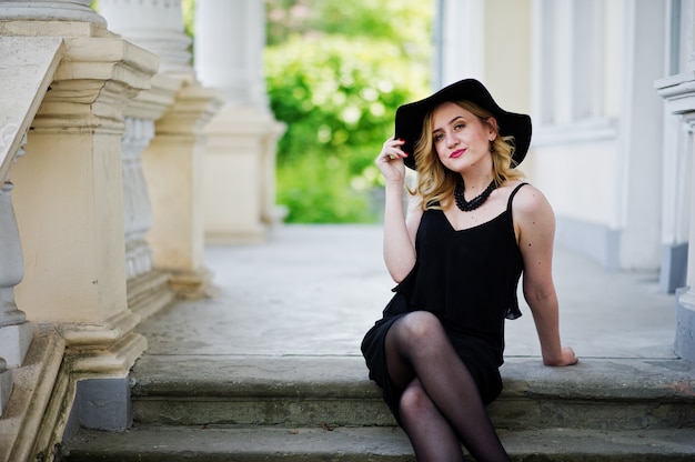 Donna bionda in abito nero, collane e cappello contro la casa d'epoca.