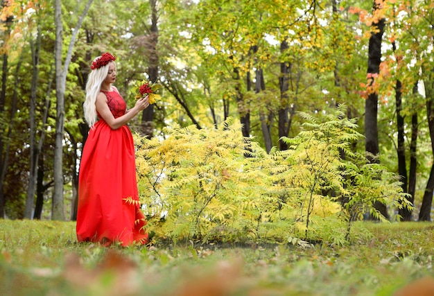 donna bionda in abito rosso nel parco autunnale con bouquet di sorbo e ghirlanda di sorbo tra i capelli
