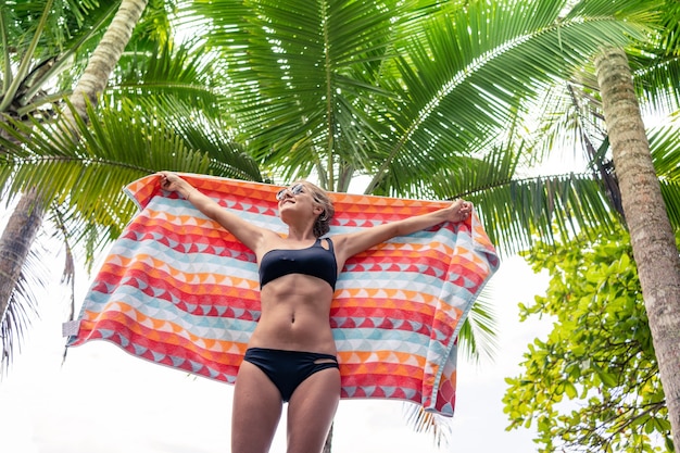 Donna bionda in bikini che sorregge un panno colorato davanti a una palma