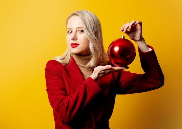 Donna bionda in cappotto rosso con pallina di Natale sulla parete gialla