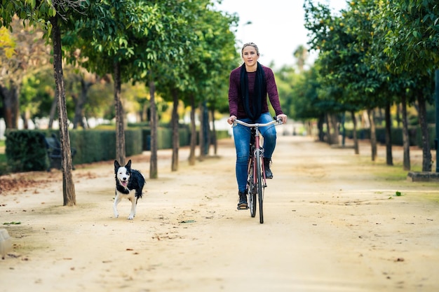 Donna bionda in denim in sella a una bici accanto a un cane che corre in un parco pubblico