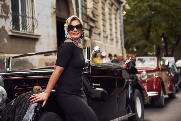 Donna bionda in occhiali da sole e in abito nero che si appoggia sulla vecchia auto d'epoca d'epoca.