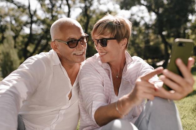 Donna bionda in occhiali da sole e vestiti rosa che tiene il telefono sorridente e posa con l'uomo con i baffi in camicia leggera all'aperto