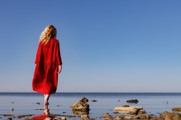 Donna bionda in un lungo abito di lino rosso in piedi in mare al mattino.