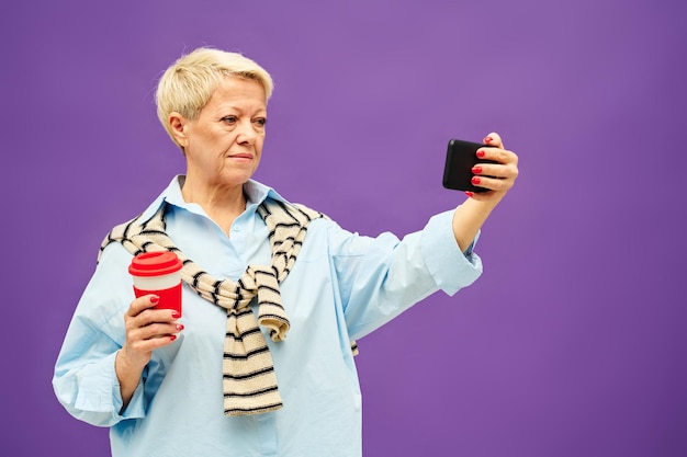 Donna bionda matura in abbigliamento casual che mangia caffè e guarda video online