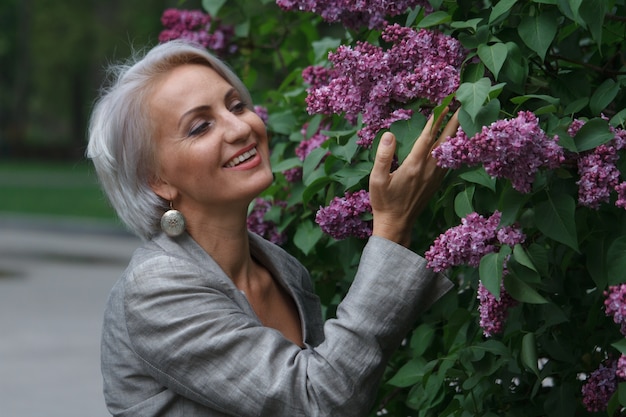 Donna bionda matura in abito grigio cammina nel giardino pubblico, ammira i fiori e sorride