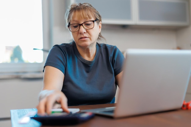 Donna bionda matura stanca freelancer che lavora da casa utilizzando il laptop seduto in cucina