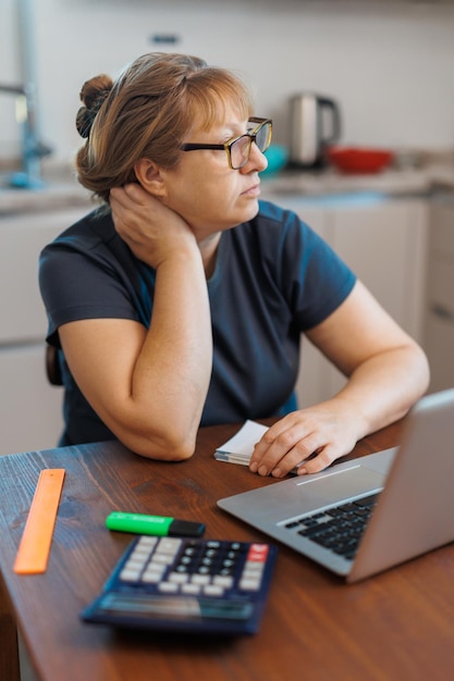 Donna bionda matura stanca freelancer che lavora da casa utilizzando il laptop seduto in cucina