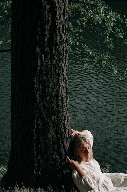 Donna bionda piuttosto sensuale in un abito bianco vintage seduto vicino a un albero in riva al fiume in una calda giornata.