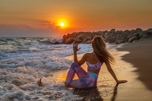 Donna bionda seduta in una posizione di torsione spinale in riva al mare a guardare il tramonto mentre si pratica yoga