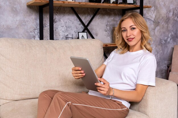 Donna bionda seduta sul divano utilizzando tablet a casa nel soggiorno
