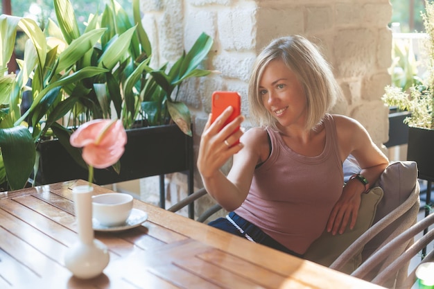 Donna bionda sorridente che fa selfie su un telefono rosso mentre è seduto in un accogliente caffè