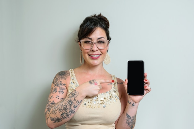 Donna brasiliana con gli occhiali, tatuata, capelli in su, sorridente e puntando il dito contro lo smatphone.