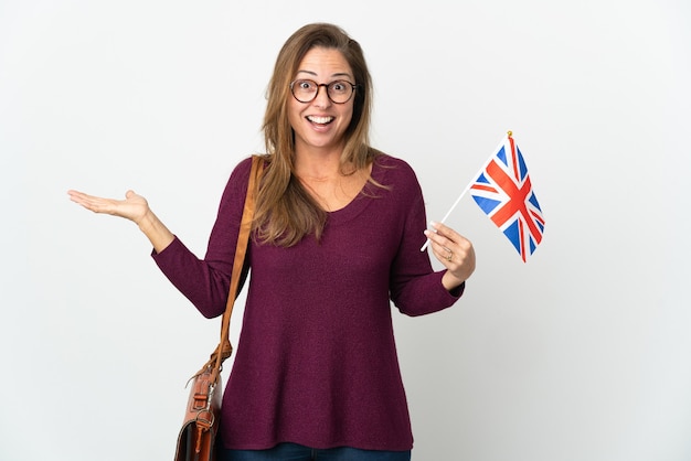 Donna brasiliana di mezza età che tiene una bandiera del Regno Unito isolata sul muro bianco con l'espressione facciale scioccata