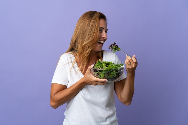 Donna brasiliana di mezza età isolata sulla parete viola che tiene una ciotola di insalata con l'espressione felice