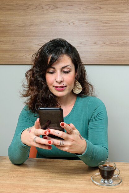 Donna brasiliana, sorridente, seduta e interagendo con il suo smartphone. Accanto a una tazza di caffè (foto verticale).