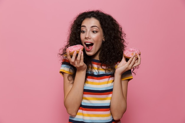 donna bruna anni '20 con i capelli ricci che gioca e mangia ciambelle isolate sul rosa