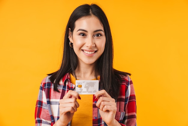 donna bruna contenta che sorride mentre tiene la carta di credito isolata sul muro giallo
