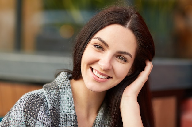 donna bruna dall'aspetto piacevole con un affascinante sorriso accattivante, avvolta in una coperta, trascorre il tempo libero nella caffetteria all'aperto