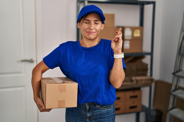 Donna bruna di mezza età che lavora indossando uniforme di consegna e berretto gesticolando dito incrociato sorridendo con speranza e occhi chiusi fortuna e concetto superstizioso