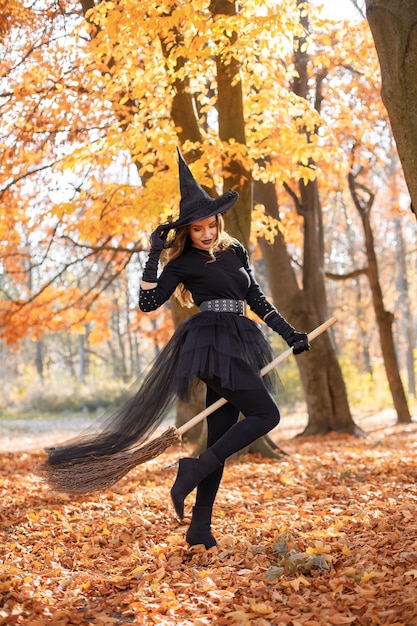 Donna bruna in costume da strega in piedi nella foresta autunnale il giorno di Halloween. Donna che indossa abiti neri e cappello a cono. Donna seduta su una scopa.