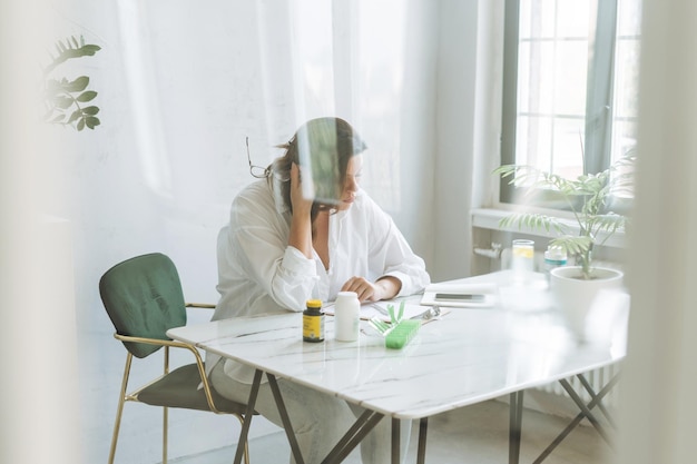Donna bruna pensante medico nutrizionista plus size in camicia bianca che lavora sul tavolo con pianta di casa nella luminosa e moderna stanza dell'ufficio