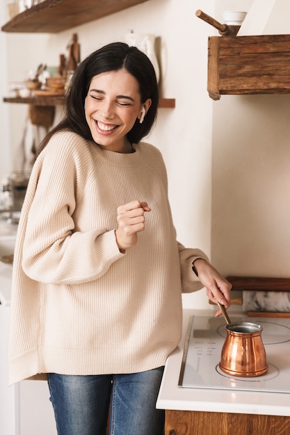 donna bruna positiva che prepara il caffè nella caffettiera turca dell'acciaio inossidabile sul piatto in cucina